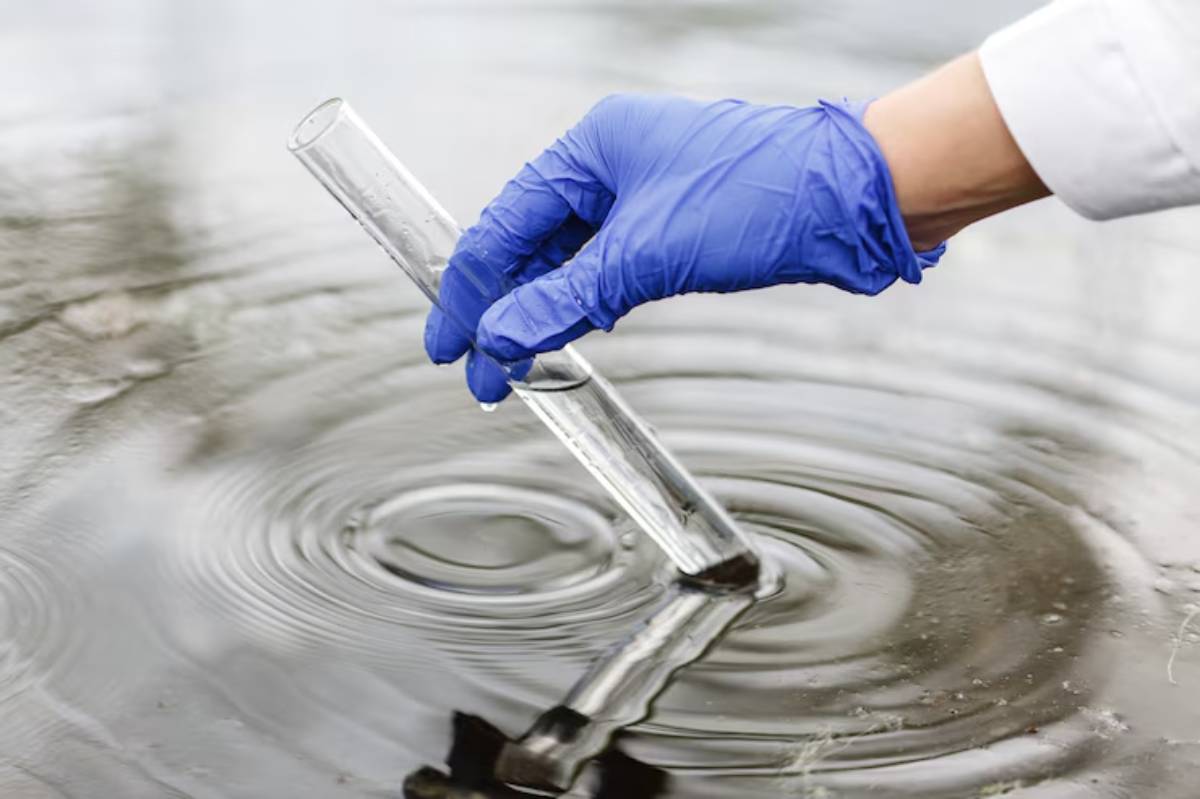 A gloved hand holds a test tube above water, creating ripples, illustrating a scientific experiment or water quality testing.